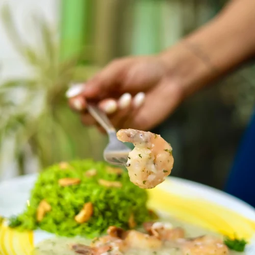 A person's hand holding a fork with a shrimp over a plate with green rice and shrimp in creamy sauce.