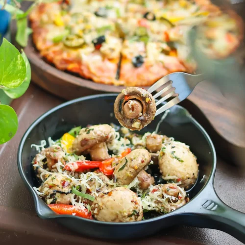 A pan of sautéed mushrooms and vegetables with a cheese-topped pizza in the background on a wooden table.