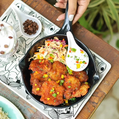 A hand holding a spoon over a pan of fried chicken with a side of coleslaw, on a patterned black and white plate.