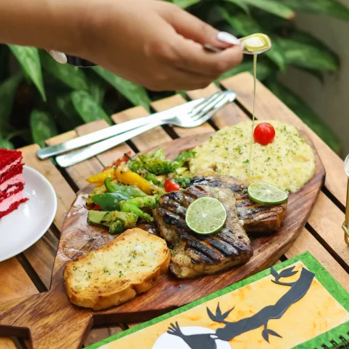 A wooden board with grilled steak, garlic bread, and vegetables, garnished with lime slices, with hands using utensils.