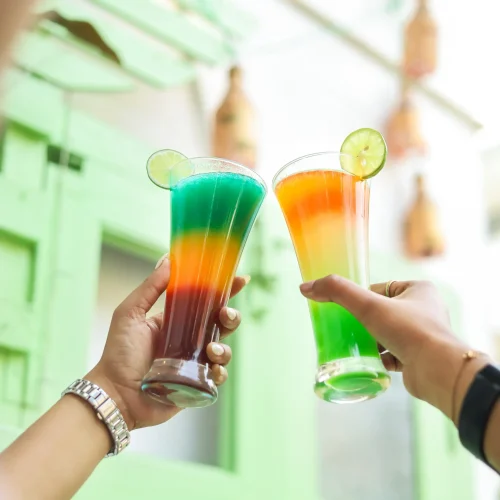 Two hands holding glasses with layered rainbow-colored drinks, garnished with lemon slices.