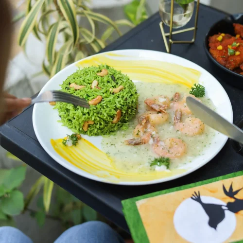 A plate with green rice and shrimp in a creamy sauce, on a black tray over a person's lap.